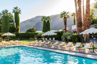 Poolside loungers and umbrellas on grass, surrounded by tall trees, mountains in background under soft sunlight.