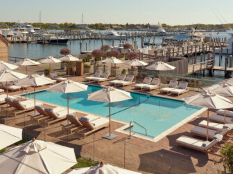 Waterfront pool area with lounge chairs and umbrellas, overlooking yachts and boats docked at scenic marina.