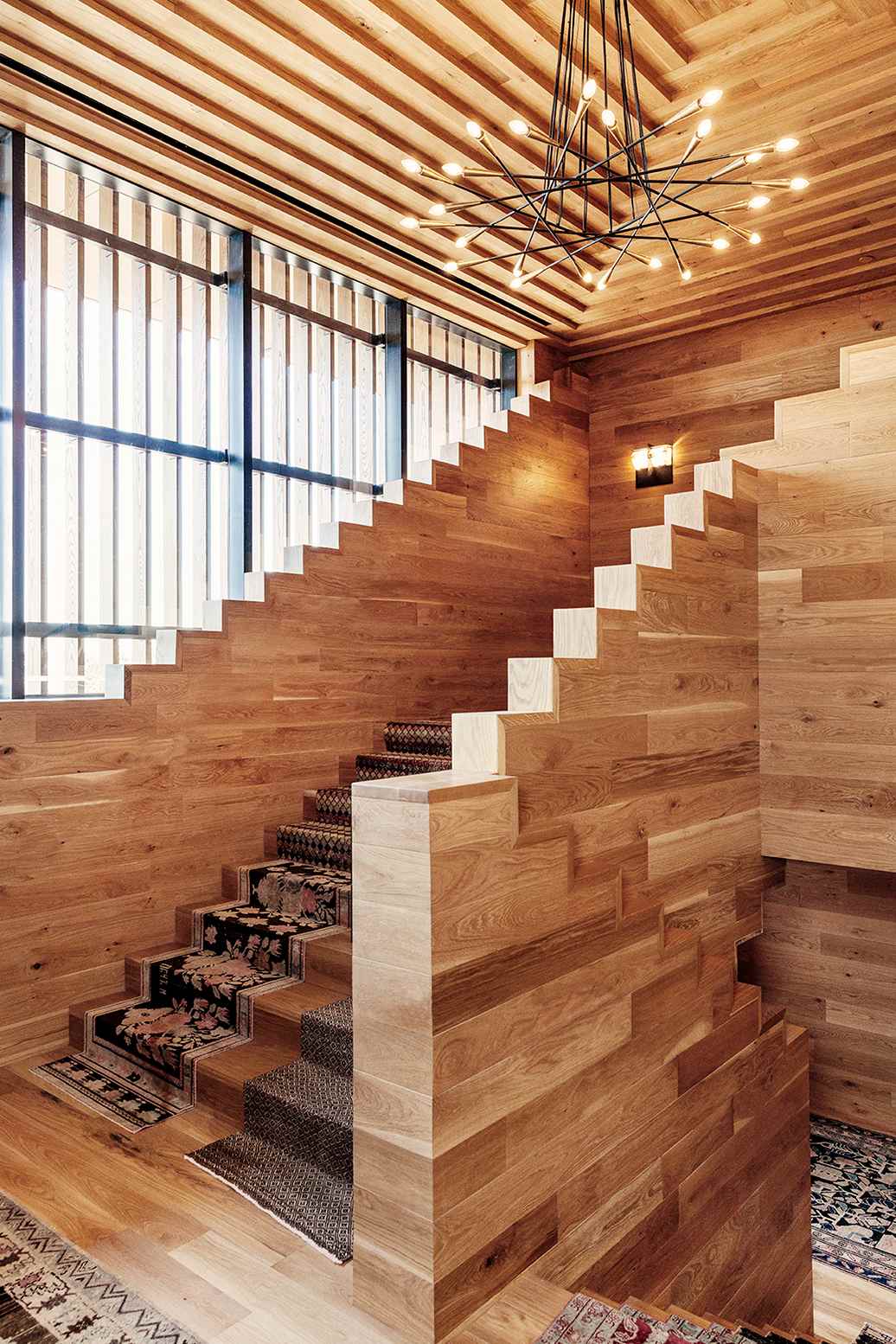 Wooden staircase with zigzag steps, slatted ceiling, patterned rugs, and chandelier in natural light.