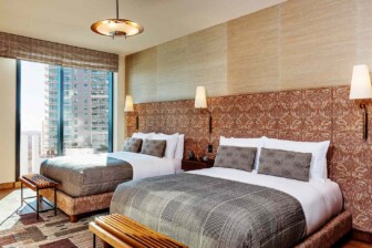 Hotel room with two beds, floral headboards, city view, and soft lighting. Neutral tones create a calm, modern retreat.