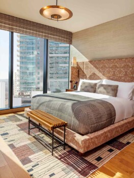 Bedroom with floor-to-ceiling windows, patterned bed, and warm tones. Sunlit, modern, city-facing comfort.
