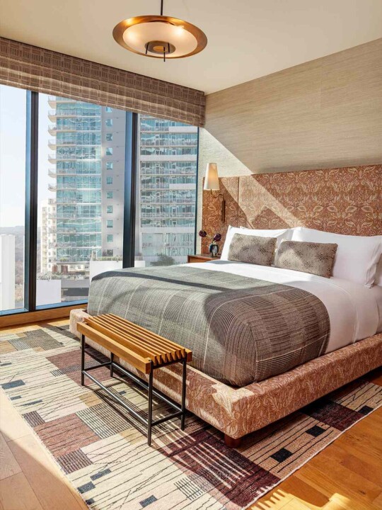 Bedroom with floor-to-ceiling windows, patterned bed, and warm tones. Sunlit, modern, city-facing comfort.