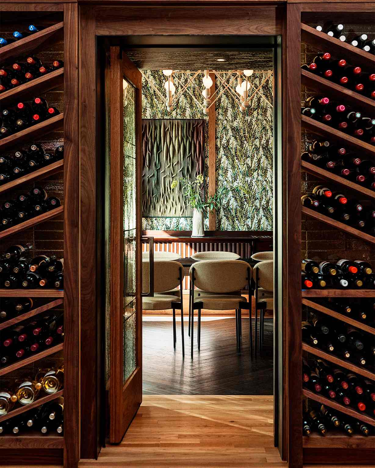 Wine-lined entry to Peacock dining room with wood table, beige chairs, and leafy wallpaper.