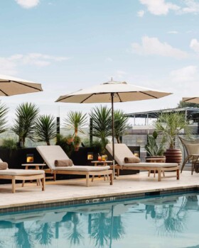 Rectangular pool with beige cushioned loungers, white umbrellas, and desert plants in urban outdoor setting.