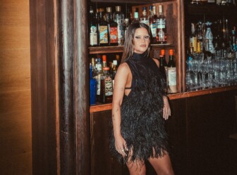 Woman in a black feather dress posing in front of bar shelves, face blurred.