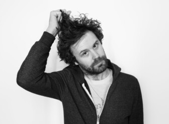 Man with curly hair tugging a handful, looking slightly puzzled against a plain background.