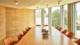 Modern conference room with long wood table, tan chairs, and large windows facing trees.