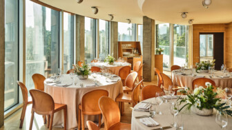 Elegant event dining room with round tables, tan chairs, and large sunlit windows.