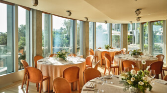 Sunlit banquet room with round tables, tan chairs, flower centerpieces, and large windows.