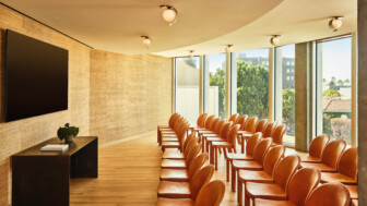 Sunlit presentation room with rows of tan chairs facing a large wall-mounted screen.