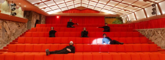 Six people sitting apart on bright red tiered seats in a modern auditorium.