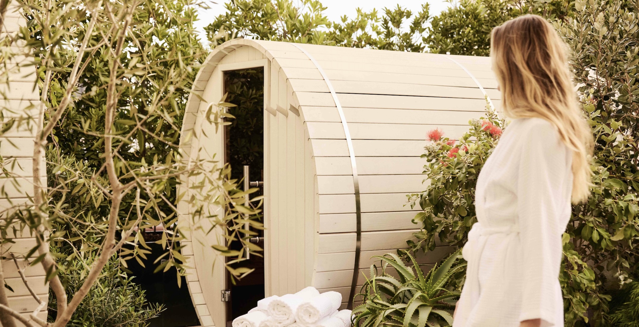 Outdoor cedar sauna surrounded by lush greenery with guest in white robe at luxury wellness retreat