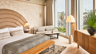 Bright hotel room with king bed, curved headboard, sofa near large windows, and wood furnishings