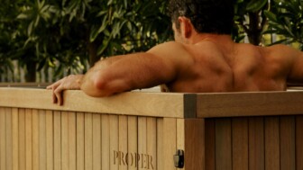 Man relaxing in a wooden outdoor soaking tub surrounded by greenery