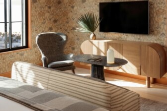 Bright hotel room with a sofa, armchair, wall-mounted TV, and small table near a window
