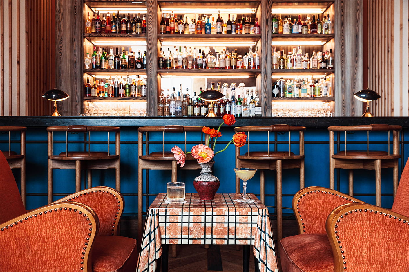 Stylish bar with backlit shelves, wooden stools, and a small table with drinks and flowers