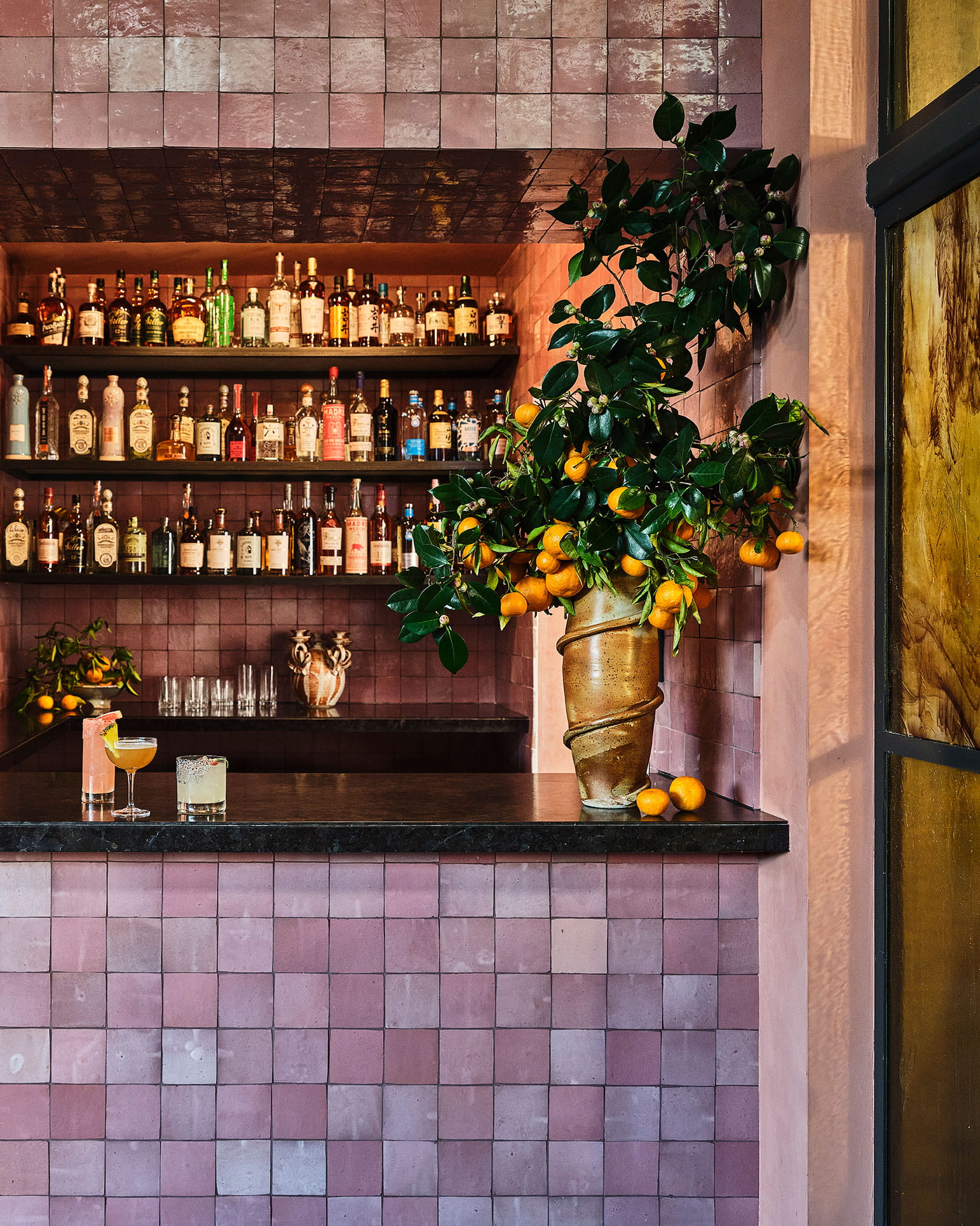 Pink-tiled bar with liquor shelves, citrus tree in a sculptural vase, and cocktails on the counter