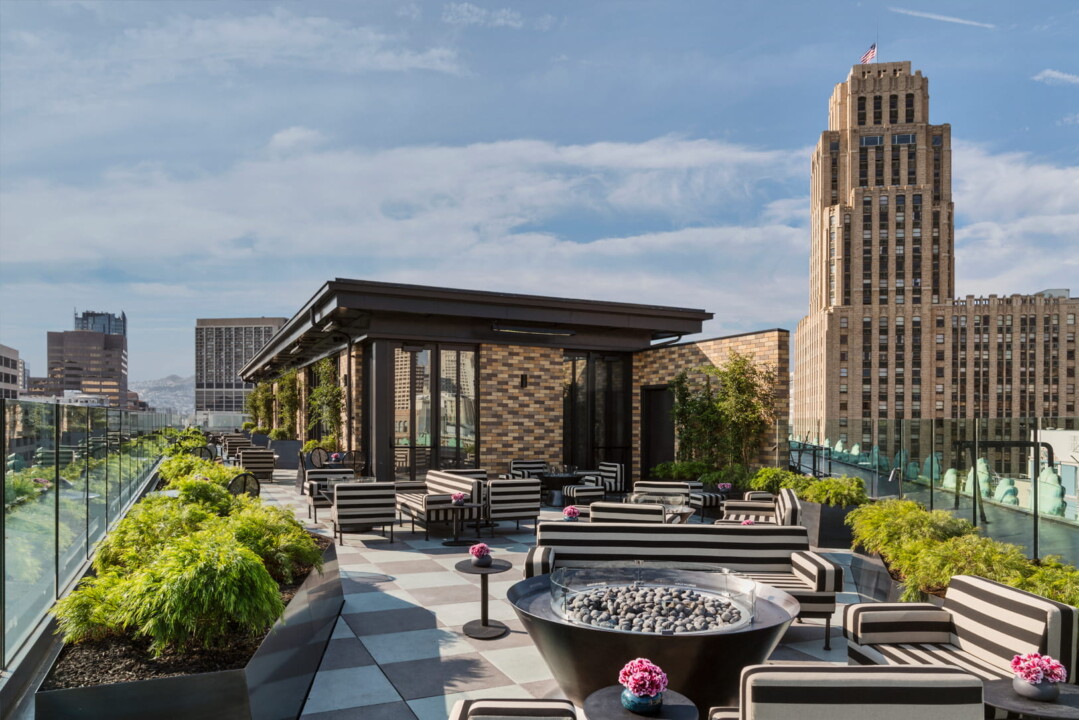 Rooftop terrace with striped lounge seating, fire pit, and city skyline views under a blue sky